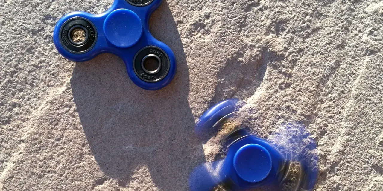 Dwa niebieskie fidget spinnery na piasku, jeden nieruchomy, drugi w ruchu, przypominają czasy, kiedy fidget spinnery były popularne.