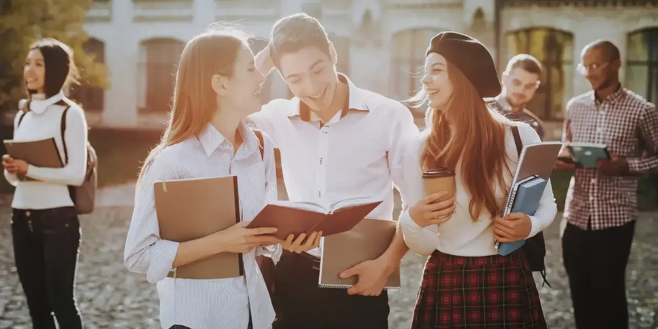 Studenci rozmawiający na kampusie, z książkami i notatkami w rękach. Wszyscy wyglądają na szczęśliwych, jakby właśnie skończyli zajęcia. To idealny obrazek dla osób, które chcą połączyć pracę z nauką, wybierając l4 a studia zaoczne.