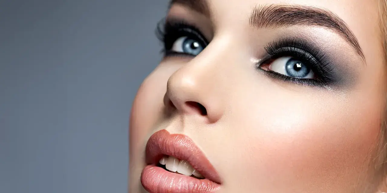 Niebieskie oczy z perfekcyjnym makijażem smokey eye, podkreślone długimi rzęsami i błyszczącymi ustami.