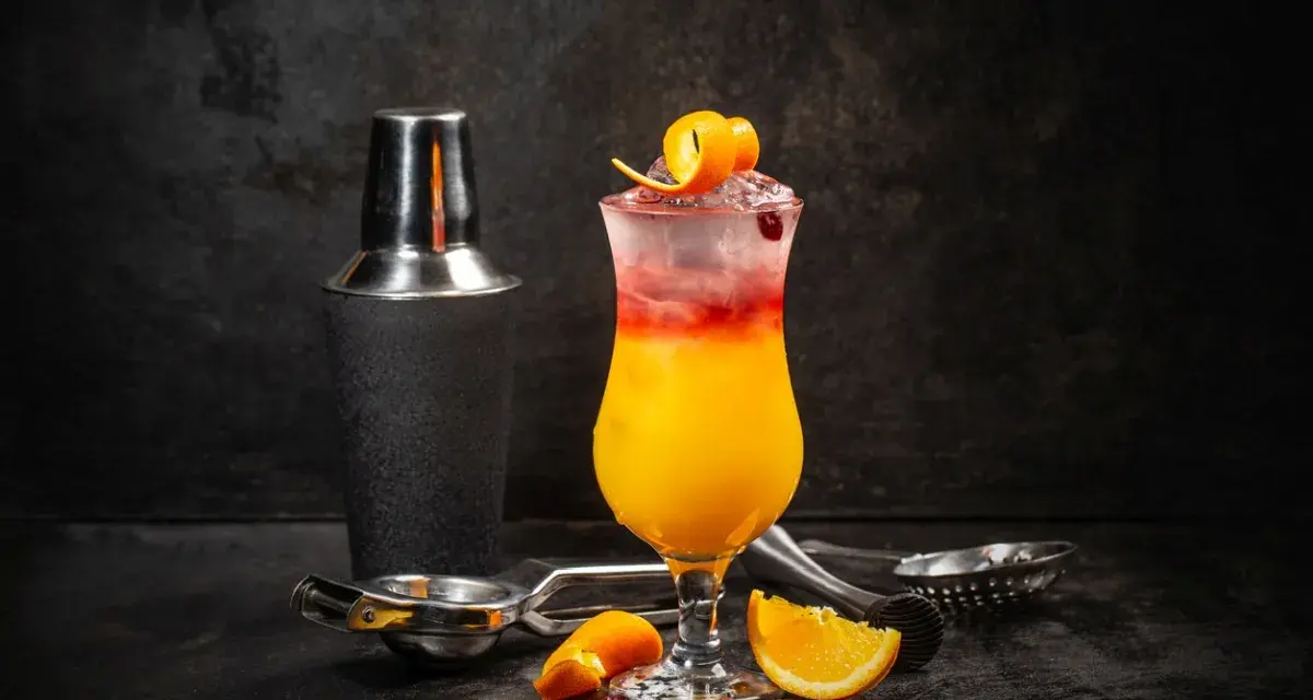 Kolorowy drink z lodem, pomarańczą i żurawiną, obok shakera. Wybór między Cosmopolitan a Sex on the Beach.