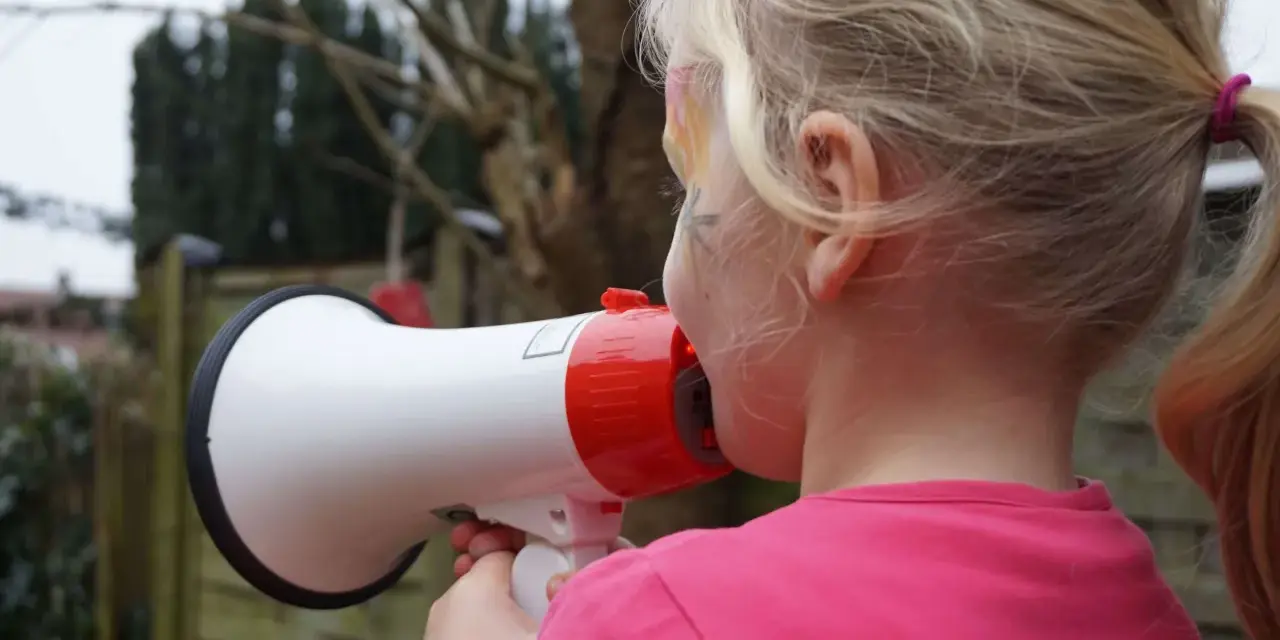 Ein Mädchen mit blonden Zöpfen hält ein Megafon und schreit hinein. Vielleicht fragt sie sich, warum sind Kinder so laut?