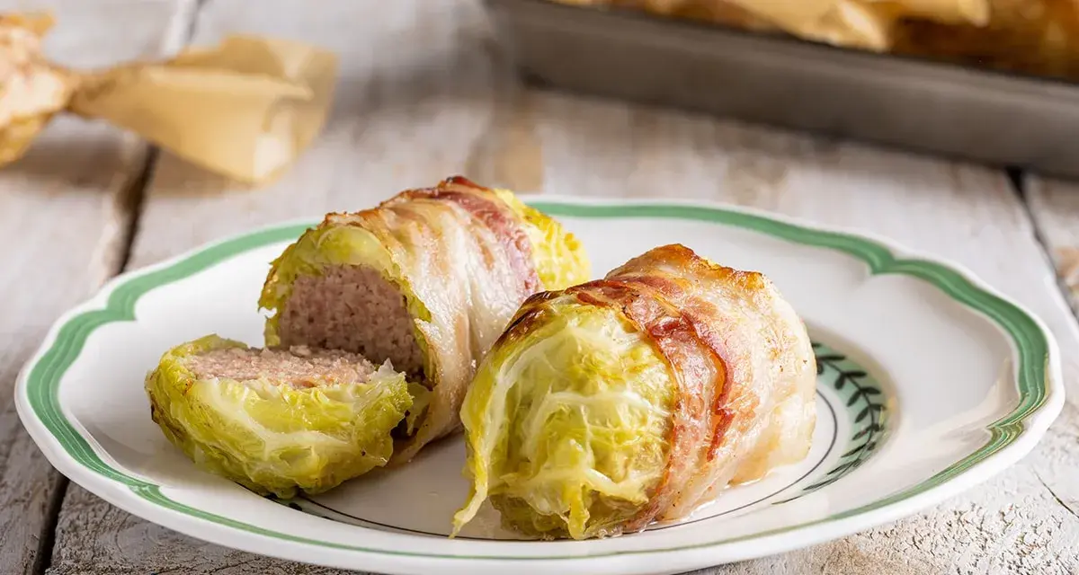 Deliziosi involtini di verza con carne, avvolti in pancetta croccante, serviti su un piatto bianco con bordo verde.