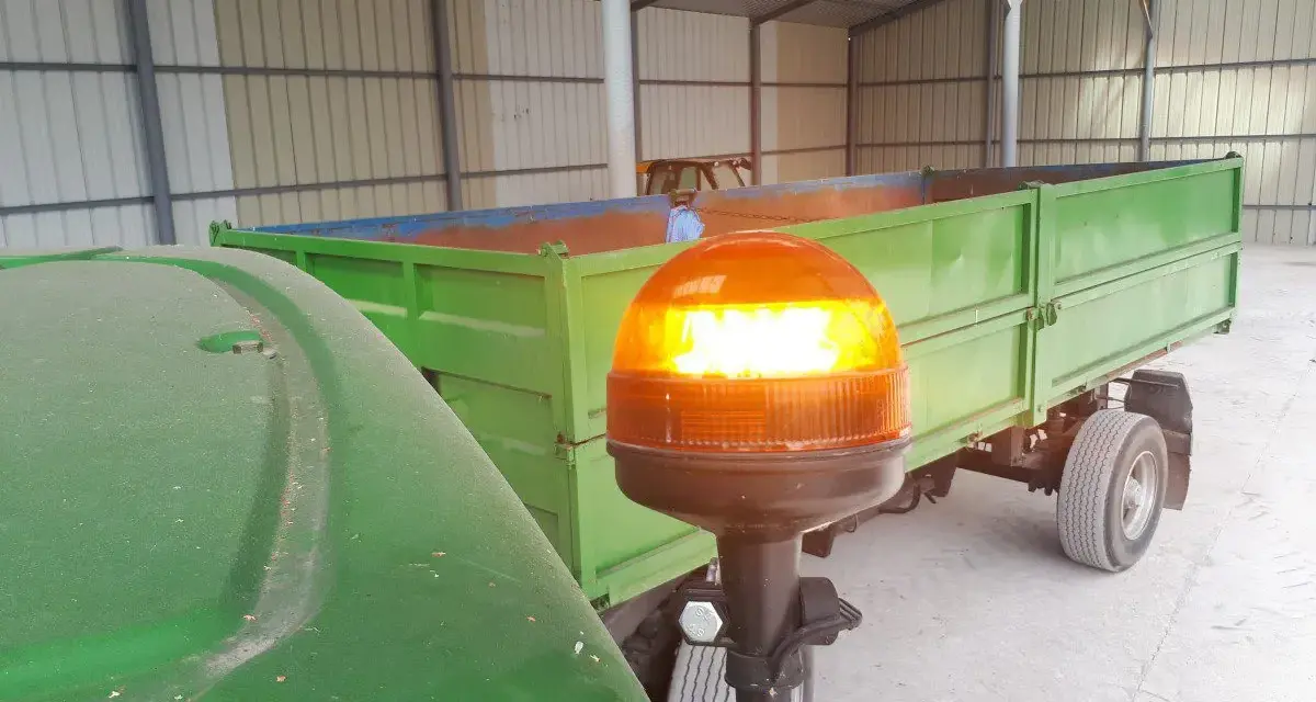 Pomarańczowa lampa ostrzegawcza na wysięgniku, przy zielonym ciągniku i przyczepie.
