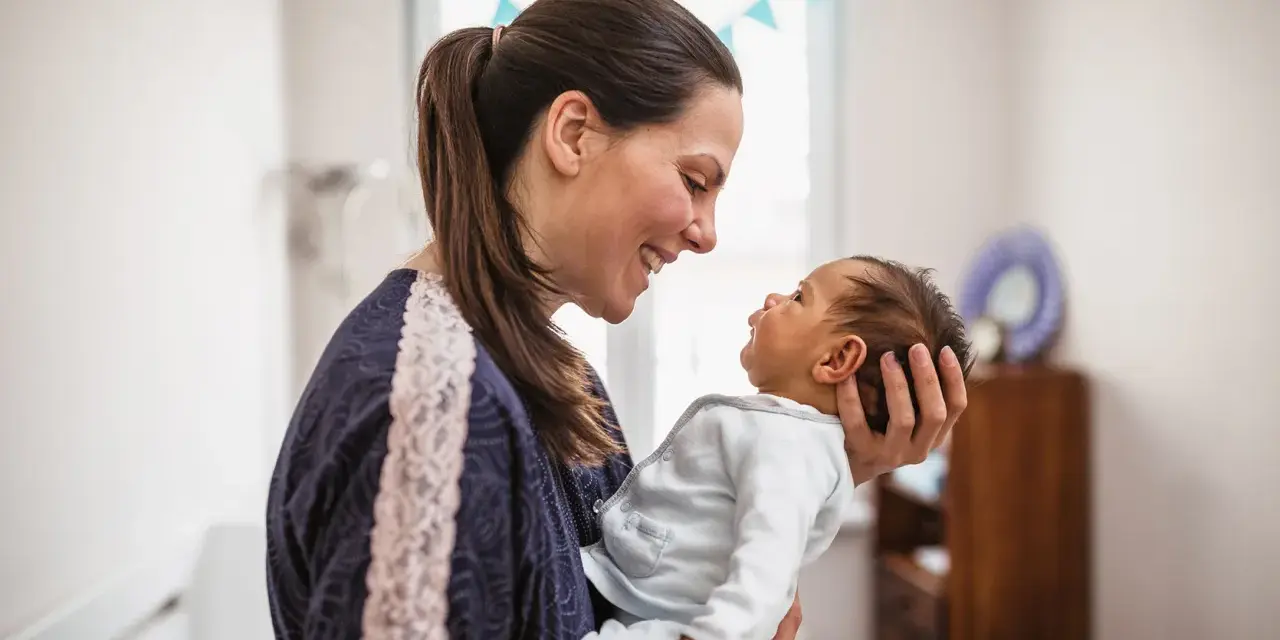 Uśmiechnięta mama trzyma na rękach noworodka. Dziecko patrzy na nią z zaciekawieniem, jakby miało zaraz kichnąć.