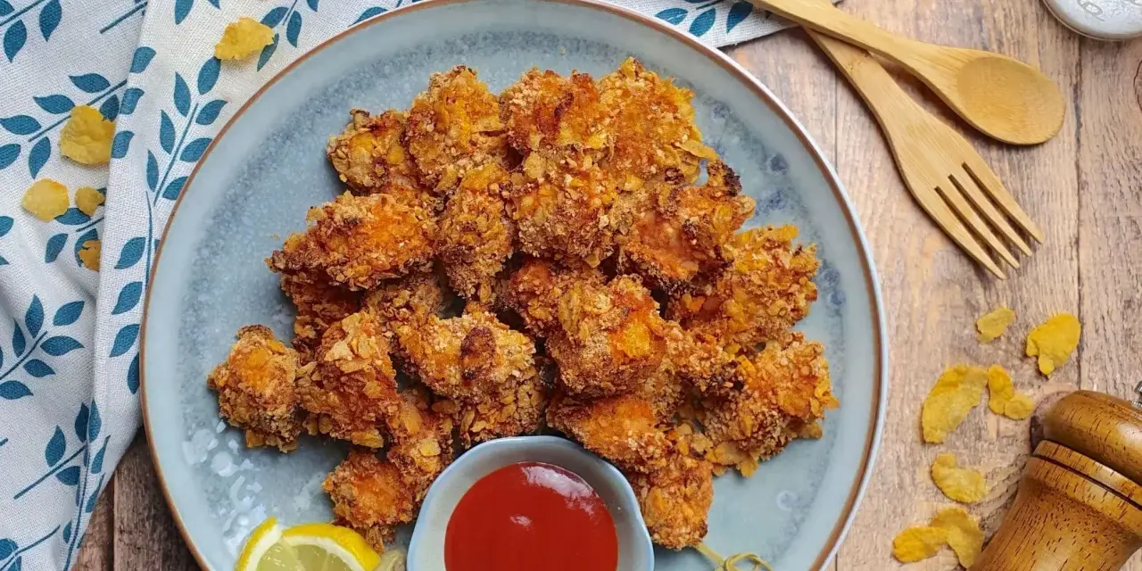 Piatto di croccanti bocconcini di pollo impanati con fiocchi di mais, serviti con ketchup e spicchi di limone.