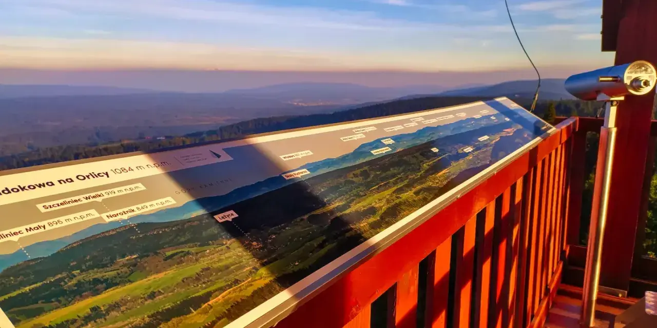 Widok z wieży widokowej na Orlicy. Na pierwszym planie panorama z opisem szczytów i dolin. W oddali łagodne wzgórza o zachodzie słońca.
