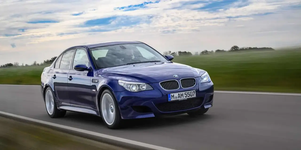 Niebieskie BMW M5 E60, ikona designu Chrisa Bangle'a, pędzi po drodze wśród zielonych pól i błękitnego nieba.