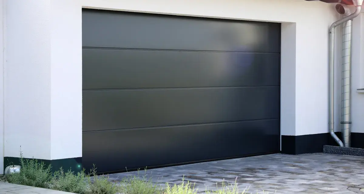 En modern, mörkgrå garageport montering med horisontella linjer. En vit fasad och grå stenläggning framför.