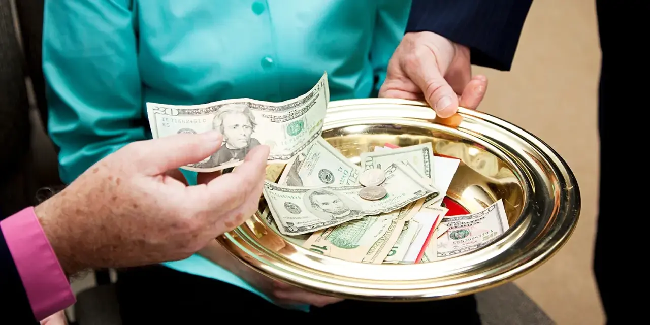 A hand places a twenty-dollar bill onto a golden tray filled with cash and coins, a gesture of offering to the poor.