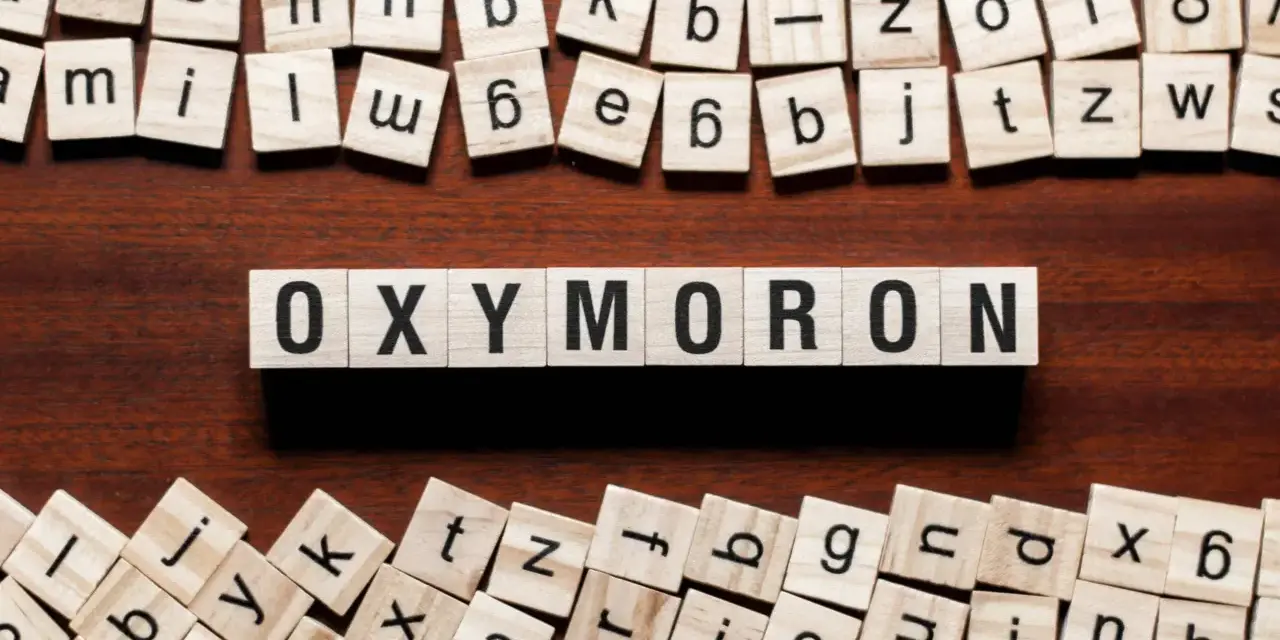 Holzbuchstaben bilden das Wort "OXYMORON". Sie sind umgeben von weiteren Buchstabenkacheln, die an Spiele wie Scrabble erinnern und Beispiele für Oxymoron-Begriffe darstellen könnten.