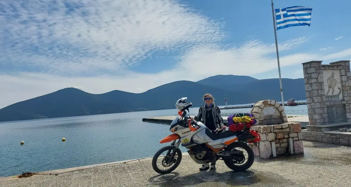 Kobieta z motocyklem nad morzem, z grecką flagą powiewającą na wietrze. Piękny dzień na podróż motocyklem nad morze.
