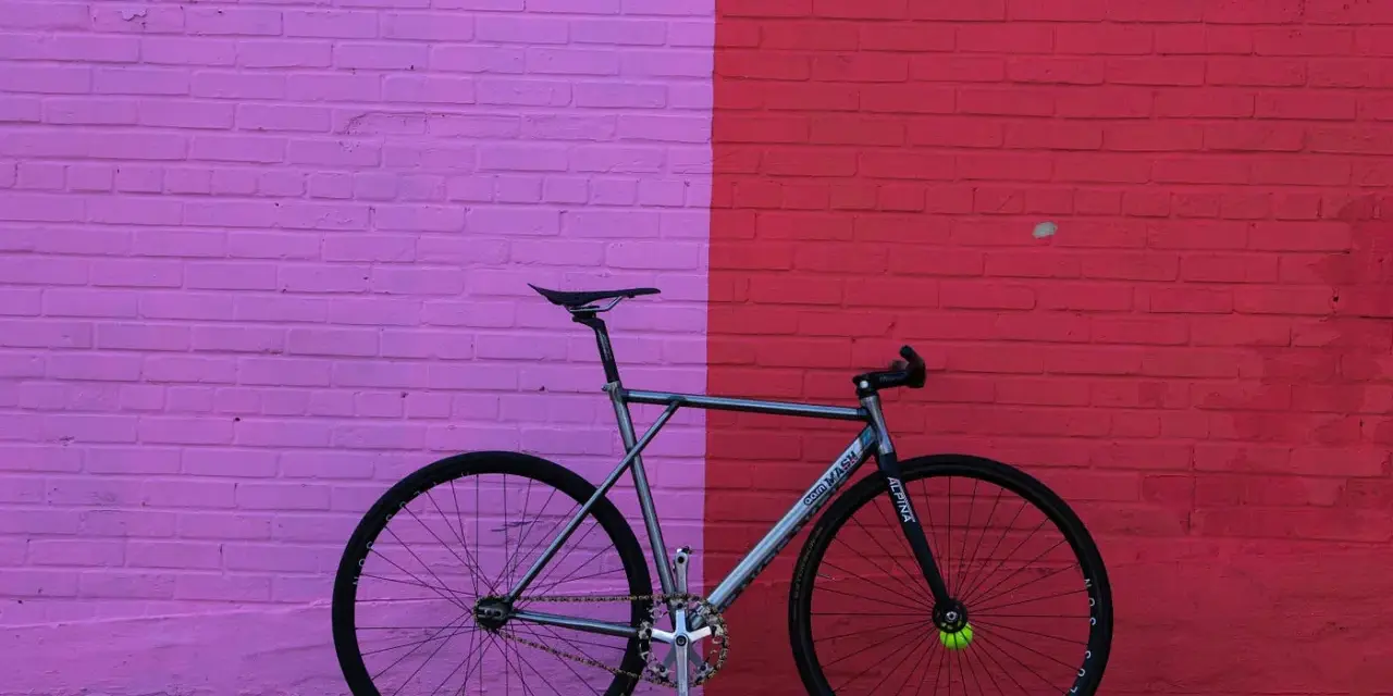 A sleek, fixed gear bike with "MASH" branding leans against a vibrant pink and red brick wall.
