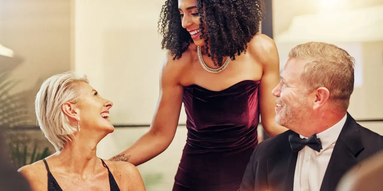 Drei Personen in eleganter Galadinner Kleidung lachen und unterhalten sich bei Kerzenlicht.