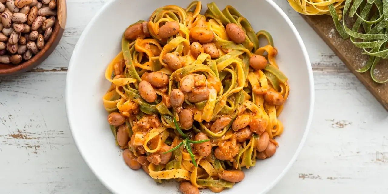 Pasta all'uovo con fagioli borlotti in una ciotola bianca, con fagioli secchi e pasta fresca colorata sullo sfondo.