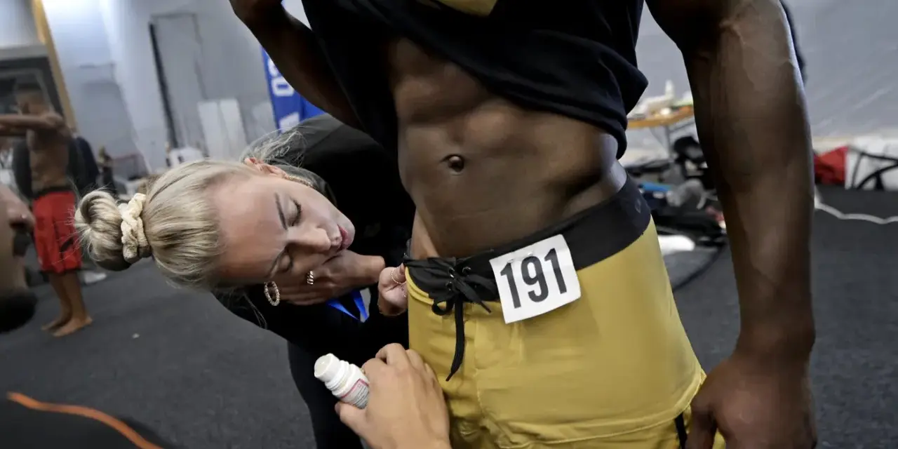 En kvinna applicerar olja på en man med en rippad kropp inför en tävling. Nummer 191 syns på hans shorts.