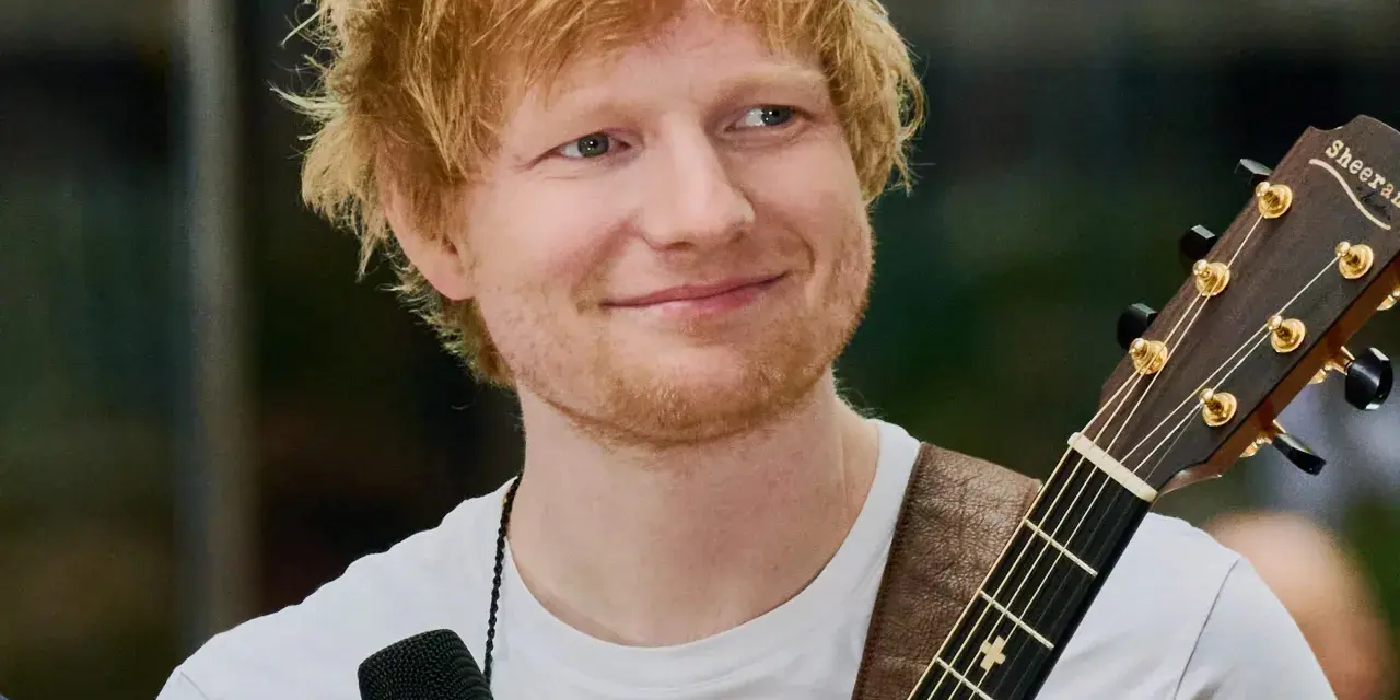 Ed Sheeran z gitarą, uśmiechnięty, w białej koszulce.