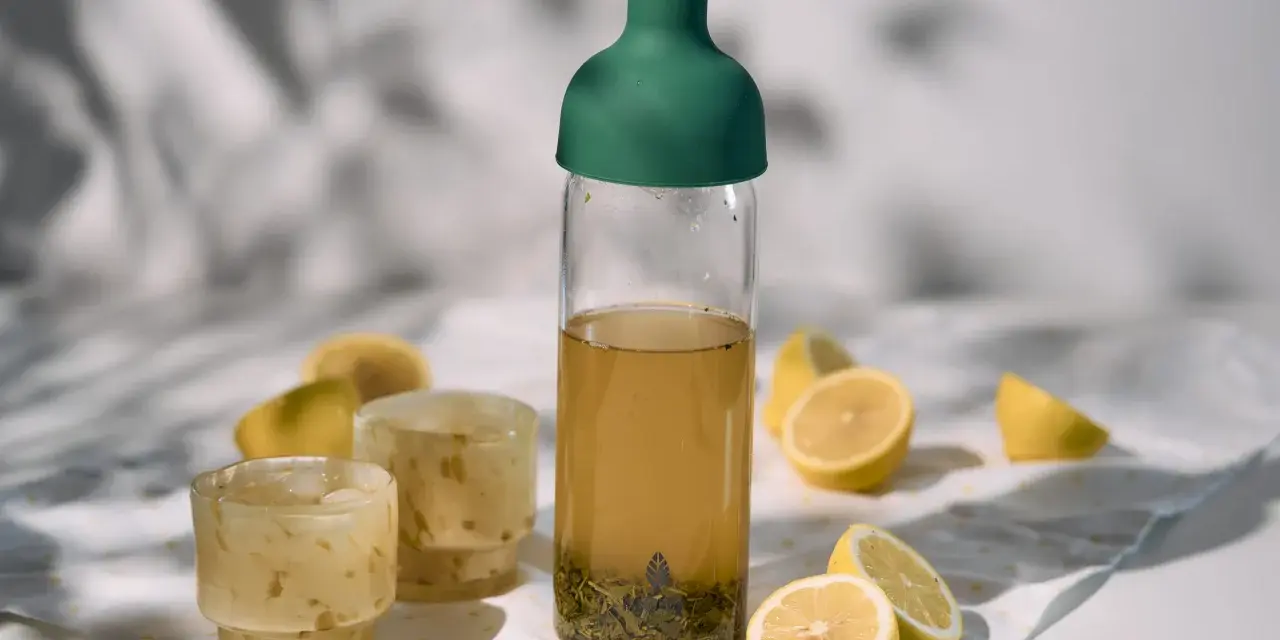 Orzeźwiający cold brew tea w butelce z zieloną zakrętką, obok szklanki z lodem i cytryny. Idealny na upalne dni.