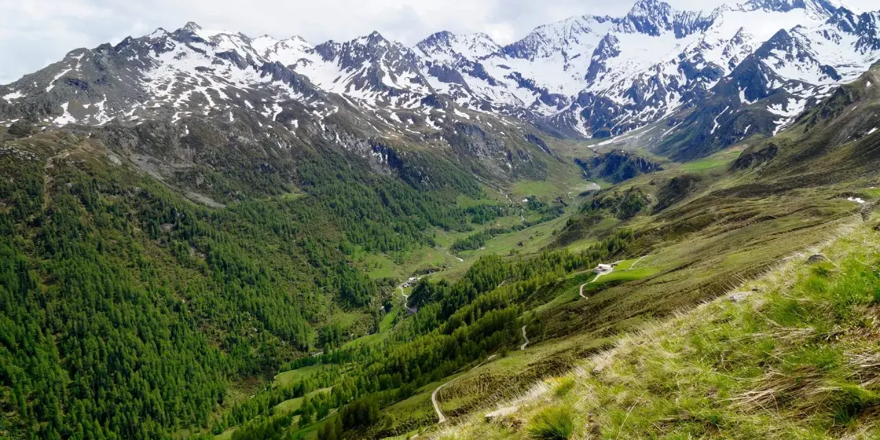 Un sentiero serpeggia su un pendio erboso verso un passo rombo in moto, con montagne innevate sullo sfondo.