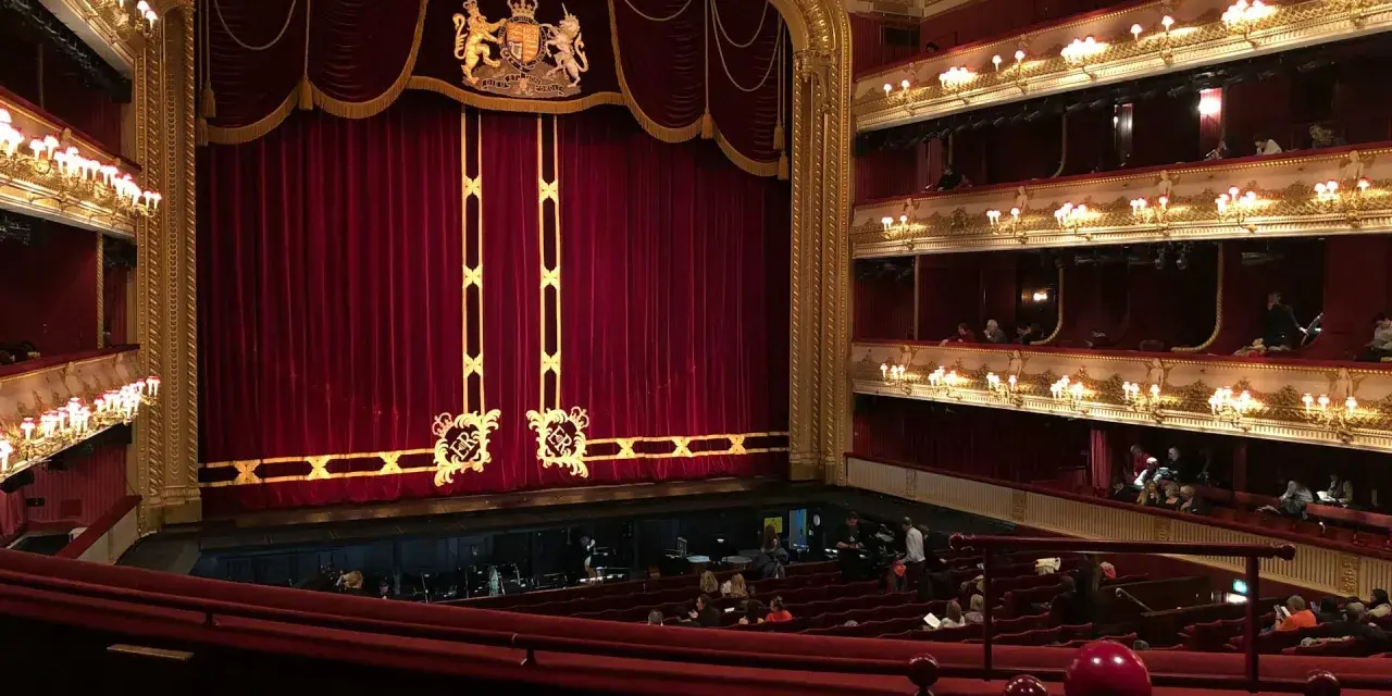 Ein prunkvolles Opernhaus mit roten Samtvorhängen und goldenen Verzierungen. Die Logen sind mit Lichtern geschmückt, und im Parkett sitzen Zuschauer.