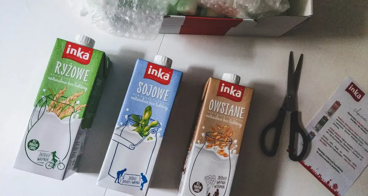 Trzy kartony napojów roślinnych Inka: ryżowy, sojowy i owsiany. Obok nożyczki i ulotka. Tak powstaje mleko owsiane.