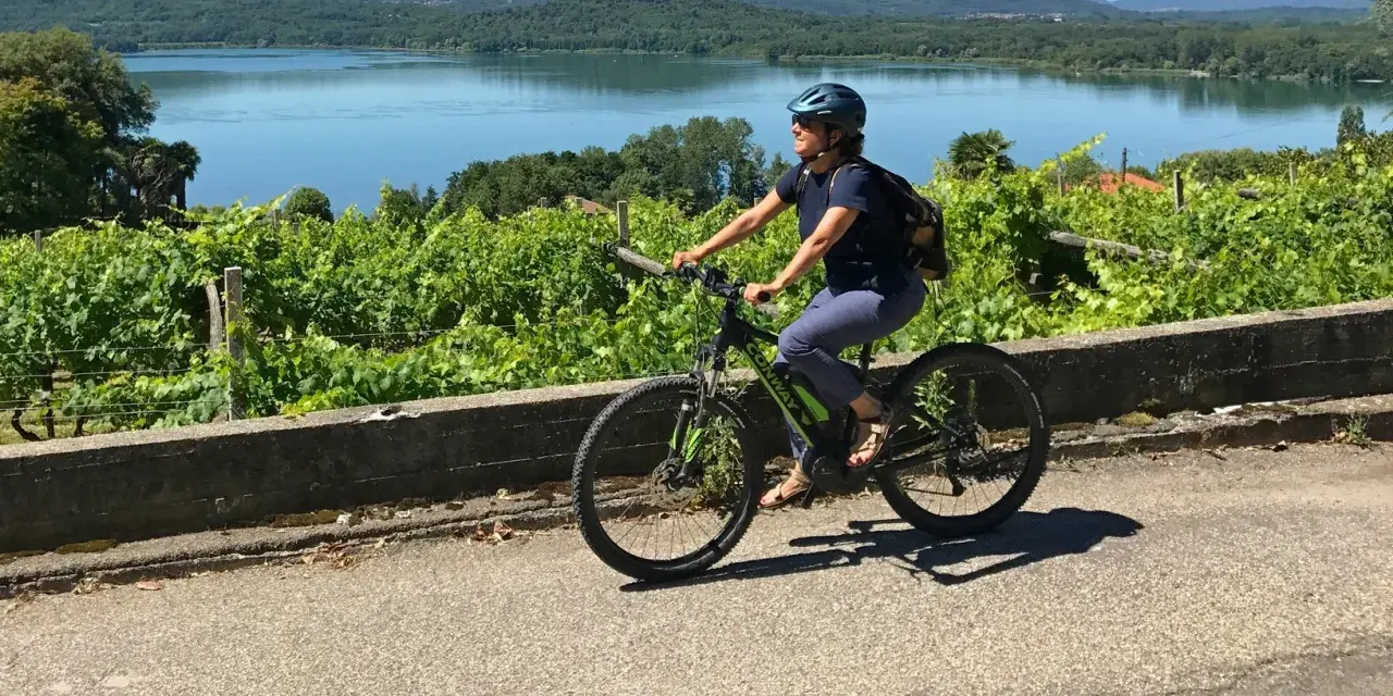 Donna in bici elettrica lungo il cammino di Oropa, tra vigneti e un lago sullo sfondo.