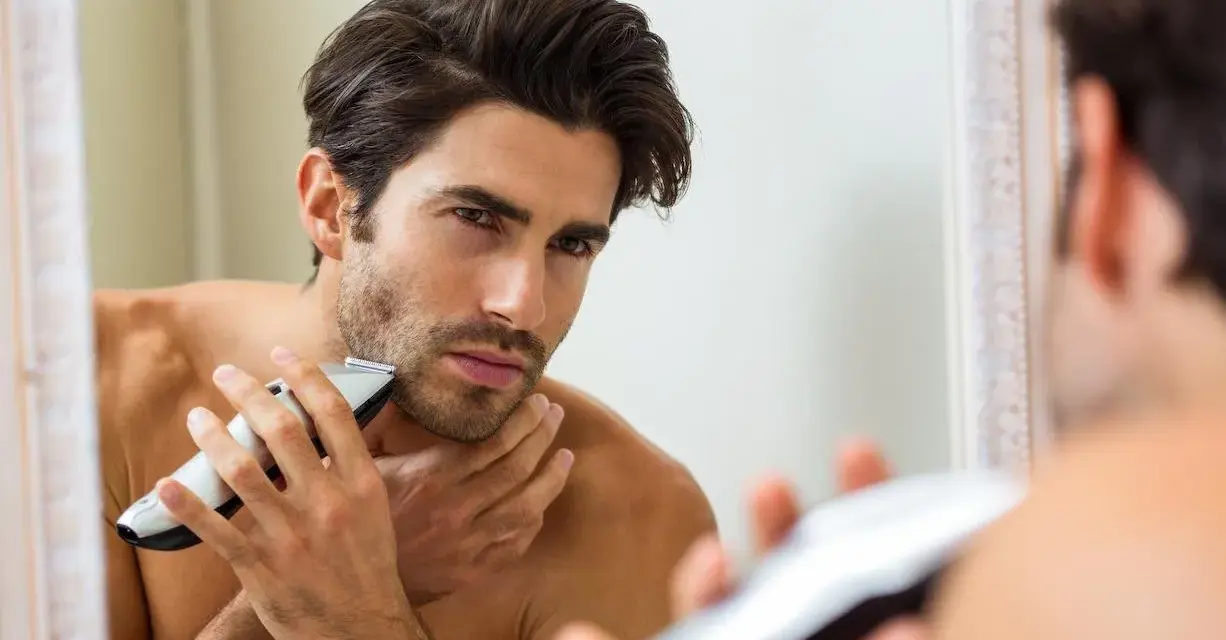 Hombre se afeita a contrapelo con una recortadora, mirándose en el espejo.