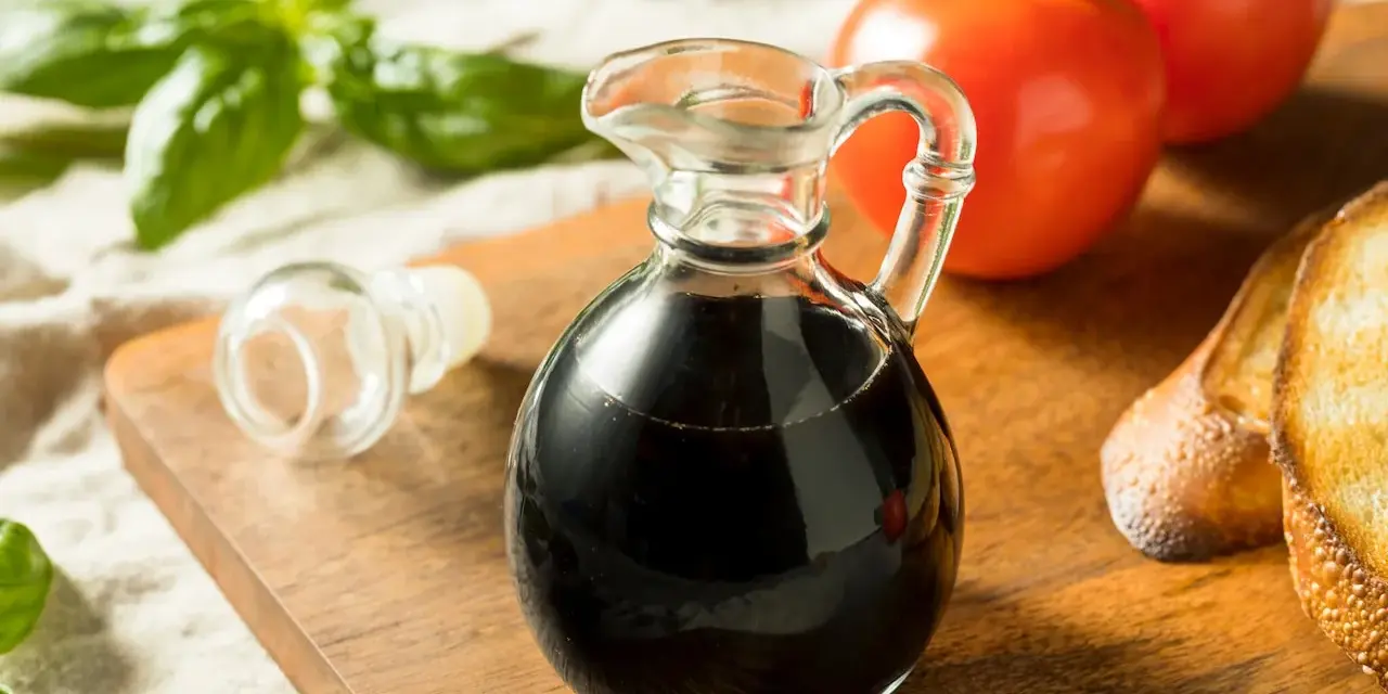 Mały dzbanuszek z ciemnym, gęstym **octem balsamicznym** obok pomidorów, bazylii i grzanek.