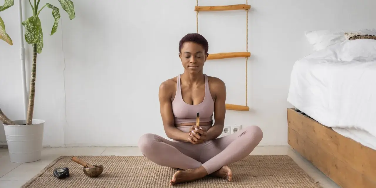 Mujer meditando en postura de loto, con los ojos cerrados y un palo de incienso en las manos. A su lado, un cuenco tibetano y una vela. Aprende como meditar para encontrar la paz interior.