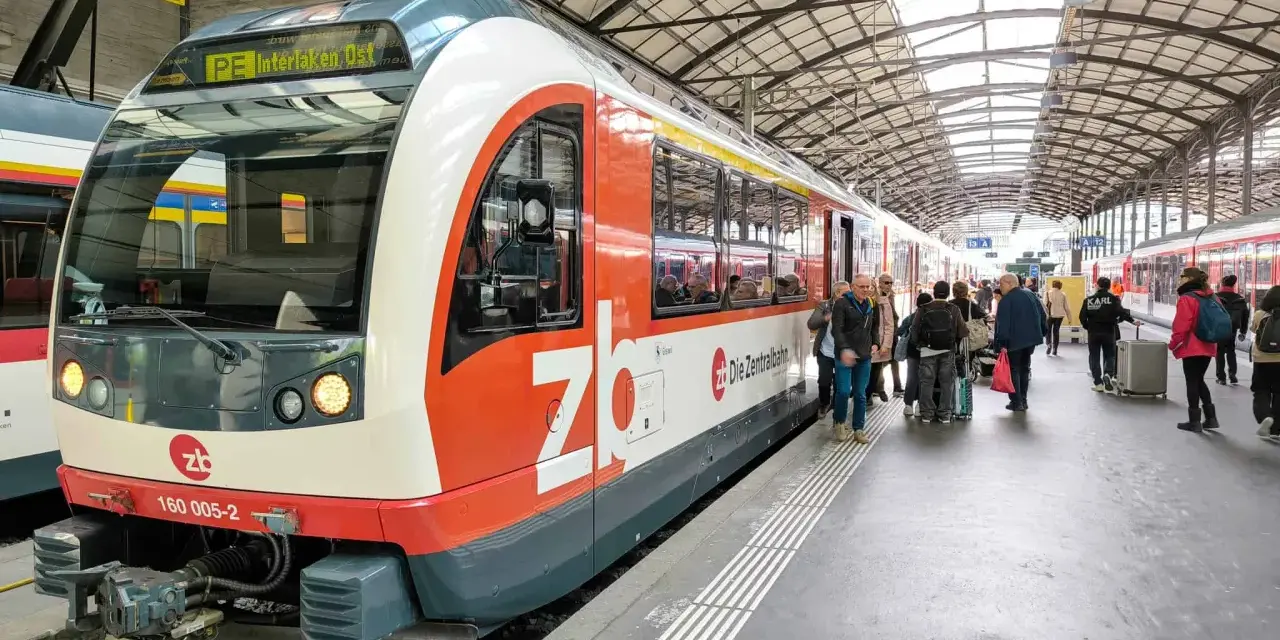 A train labeled "PE Interlaken, Ost" waits at the station, ready for its journey on the Lucerne to Interlaken Golden Pass line. Passengers gather on the platform.