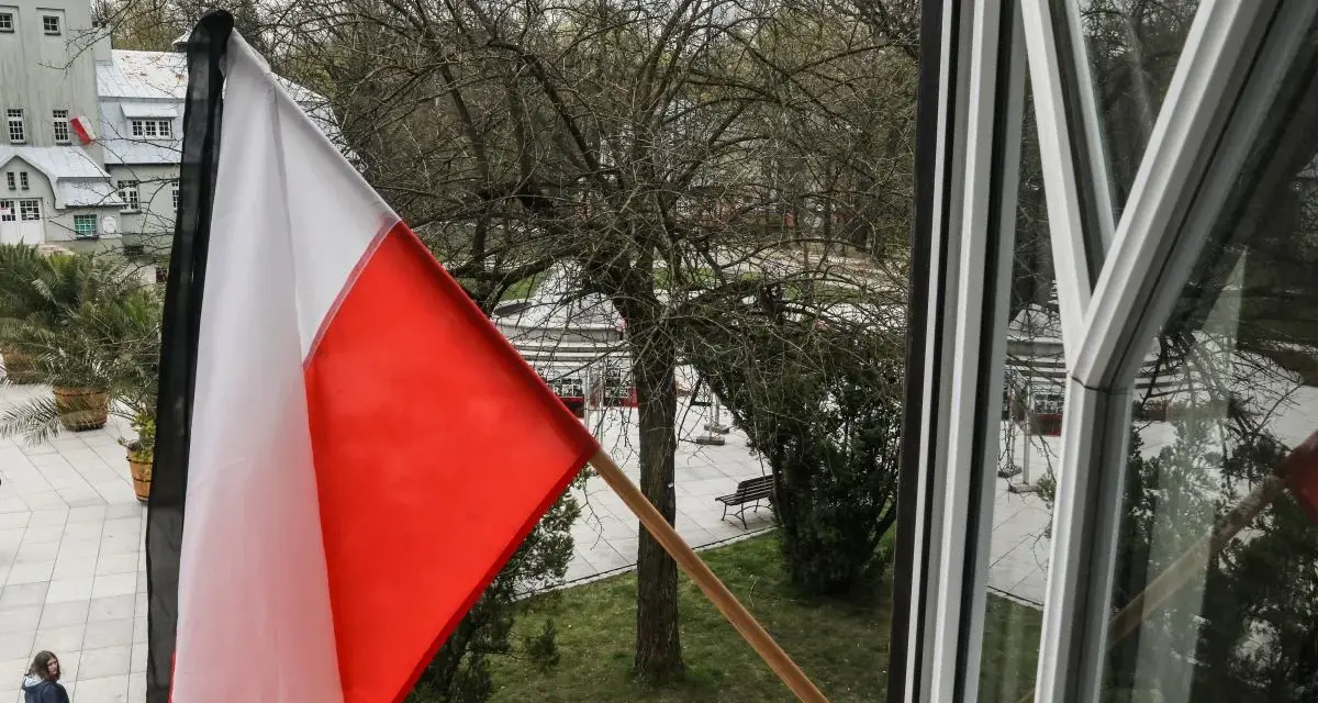 Flaga państwowa w biało-czerwonych barwach powiewa na wietrze. W tle widać drzewa i budynki.