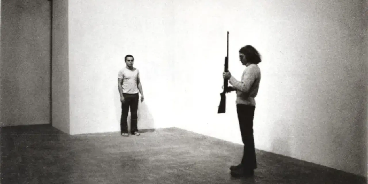 Chris Burden, dans une performance tendue, tient un fusil. Un homme se tient à distance, le regard fixé.