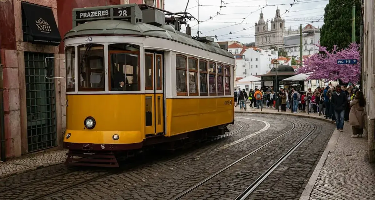 Żółty tramwaj 28 na brukowanej ulicy w Lizbonie, z ludźmi czekającymi na przystanku i kościołem w tle.