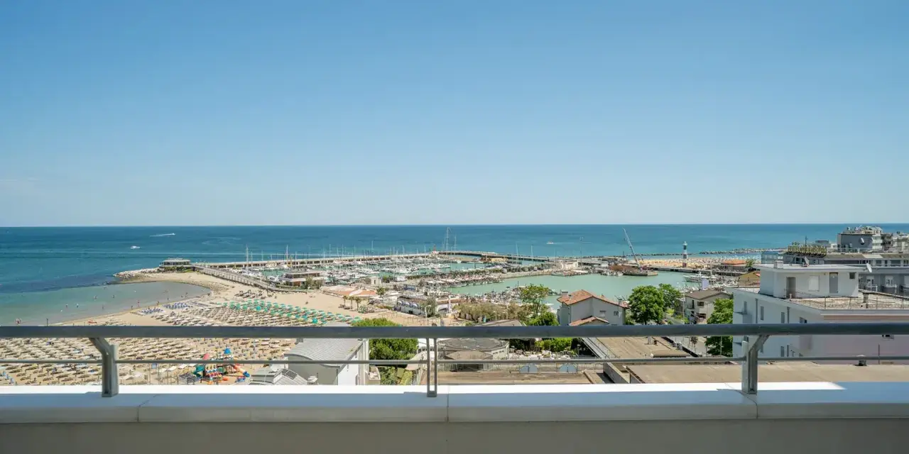 Vista dal balcone dell'hotel Acropolis Cattolica: spiaggia, porto turistico e mare azzurro sotto un cielo limpido.