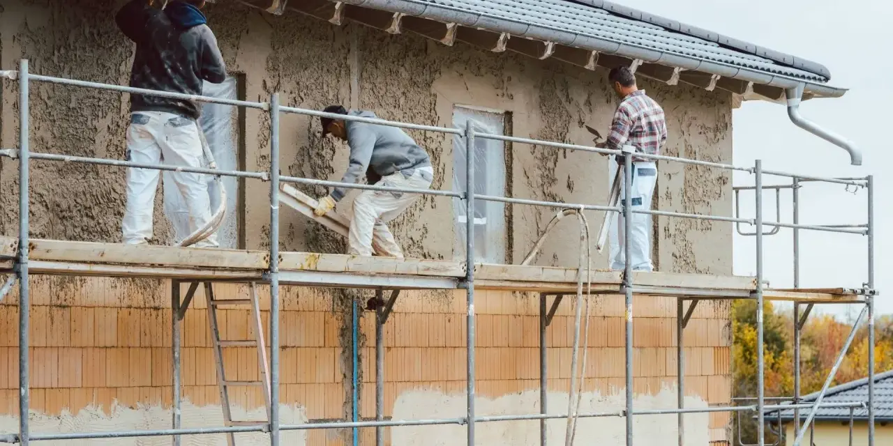 Pracownicy na rusztowaniu wykonują tynkowanie elewacji domu, nakładając zaprawę na ściany.