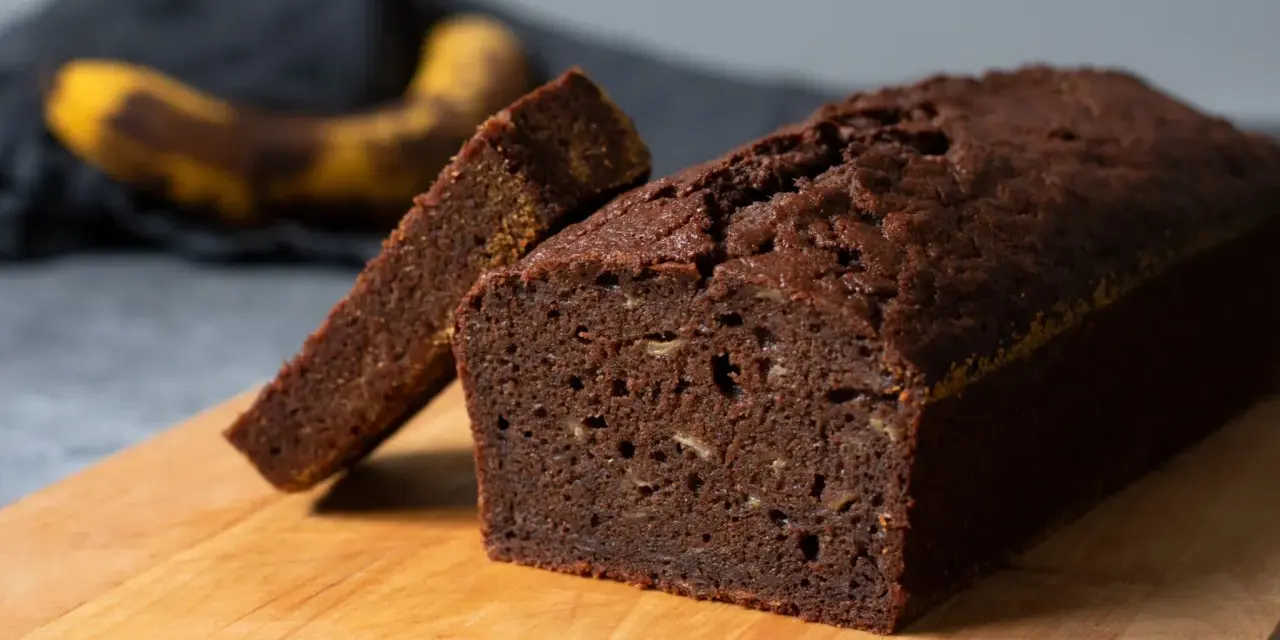 Chokladig banankaka, perfekt för ett enkelt banankaka recept. En skiva är uppskuren och en övermogen banan ligger i bakgrunden.