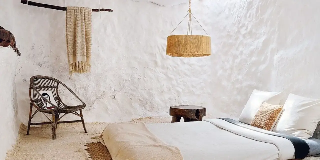 Habitación rústica con paredes de encalar, cama baja, silla de mimbre y manta de lana.