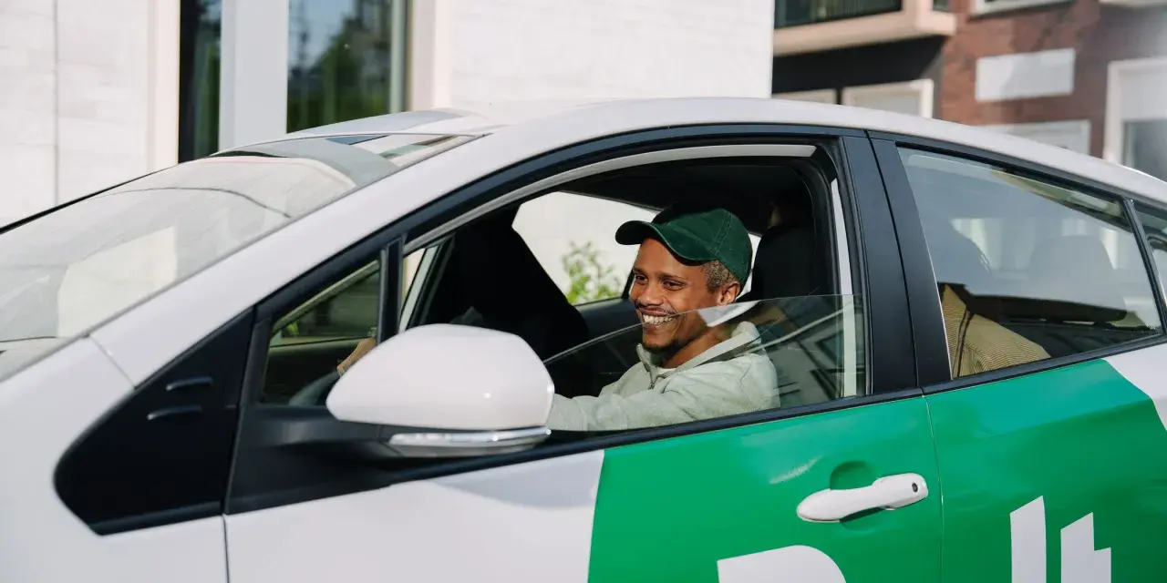 Lächelnder Mann mit grüner Kappe am Steuer eines weißen Autos mit grünen Bolt-Aufklebern. Er wird bald ein Bolt-Fahrer sein.