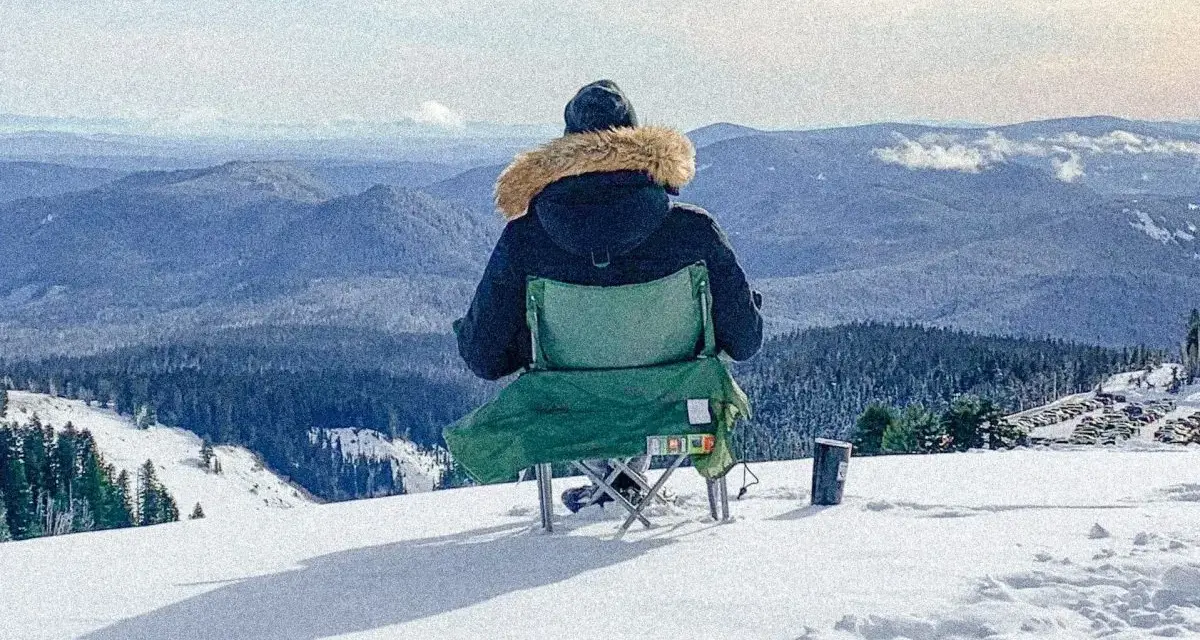 Persona sentada en silla plegable en la nieve, mirando un paisaje montañoso cubierto de árboles.