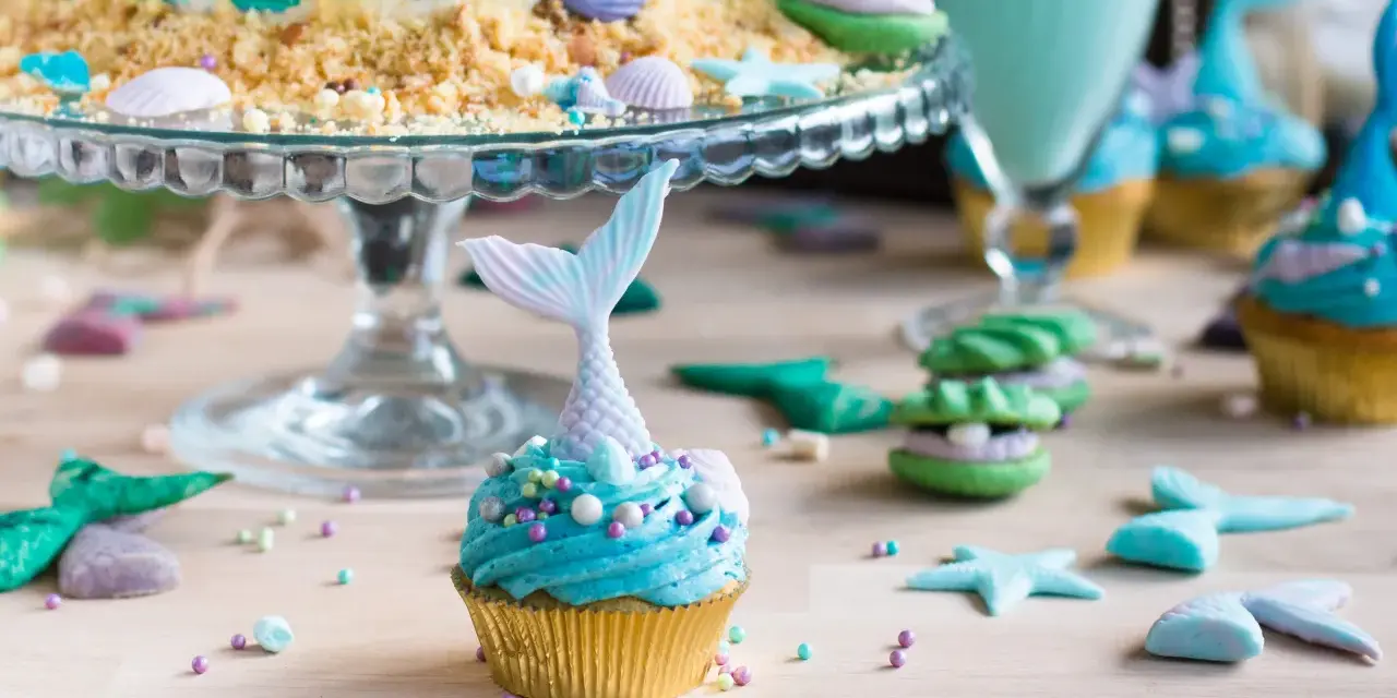 Meerjungfrau Muffins mit blauer Glasur und Schwanz-Dekorationen, umgeben von Muscheln und Seesternen.