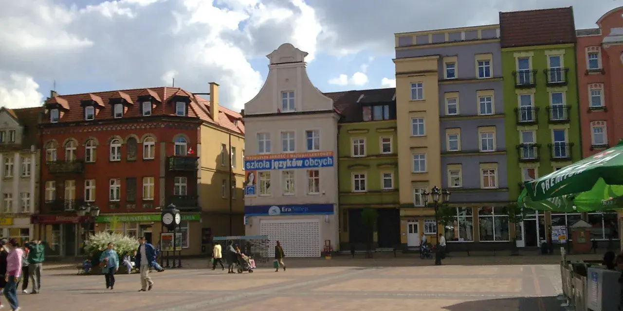 Rynek w Chojnicach z kolorowymi kamienicami, w tym ratusz. Ludzie spacerują po brukowanym placu.