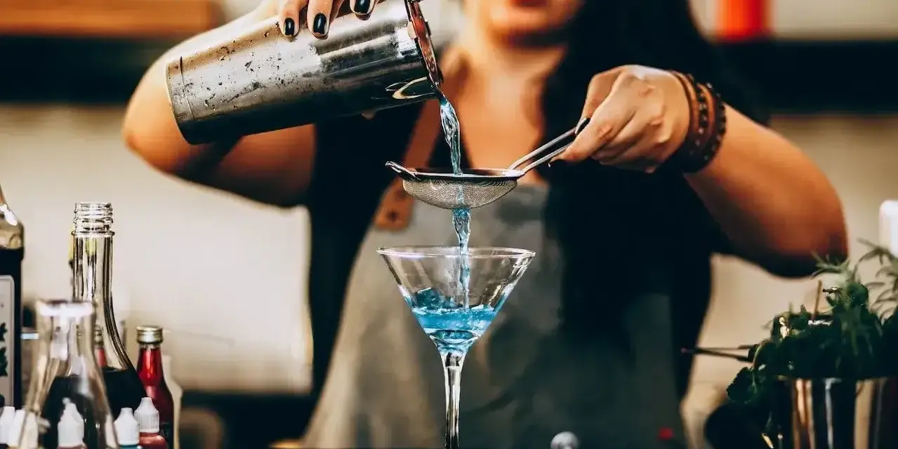 Barman przygotowuje drinka, przelewając niebieski płyn przez sitko do kieliszka. W tle widać butelki z różnymi nazwami drinków alkoholowych.