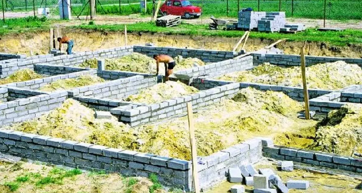 Budowa fundamentu z bloczków betonowych. Dwóch robotników pracuje przy wznoszeniu ścian fundamentowych.
