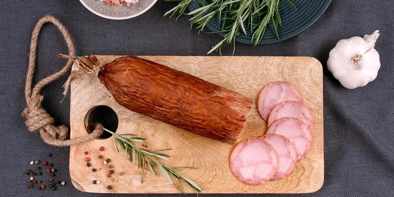 Kiełbasa żywiecka na desce, obok gałązka rozmarynu, czosnek i kolorowy pieprz. Kilka plastrów już pokrojonych.