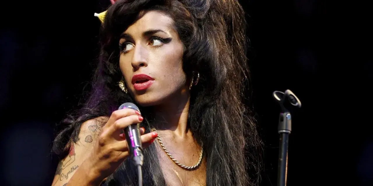 Amy Winehouse z charakterystyczną fryzurą i makijażem, śpiewająca do mikrofonu. Jej styl przypomina legendy z klubu 27, a jej muzyka wciąż żyje w sercach fanów w Polsce.