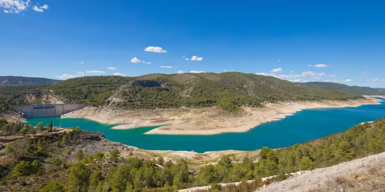 El pantano de Sacedón, con su agua turquesa, se extiende entre colinas verdes bajo un cielo azul.
