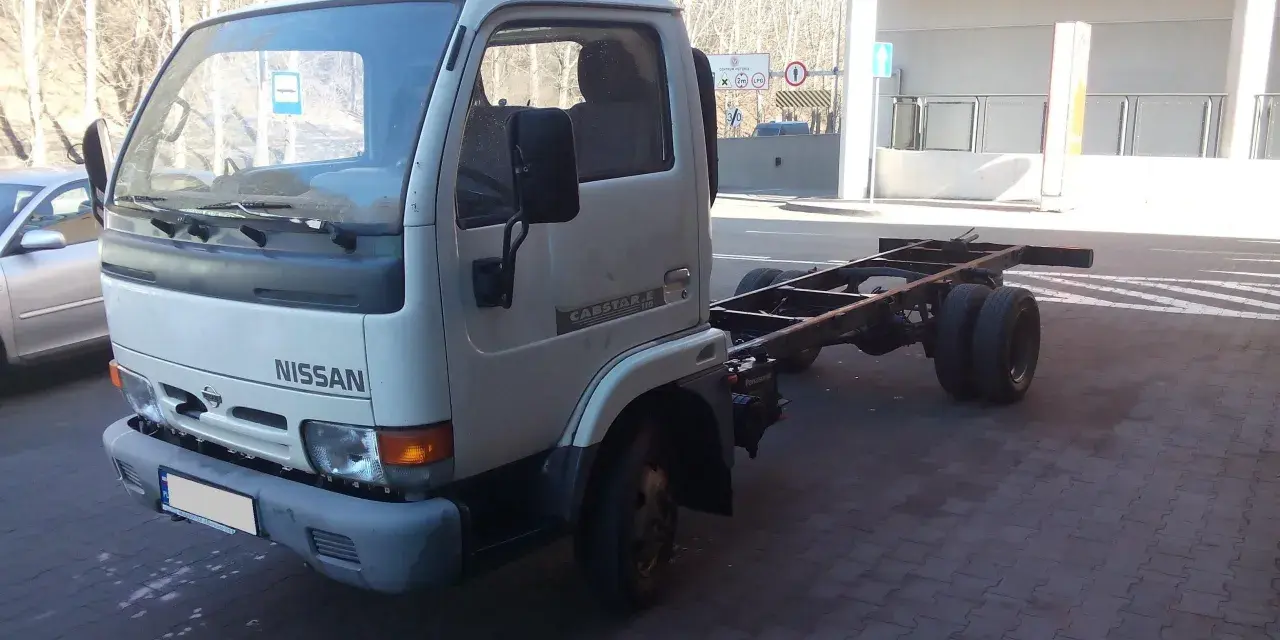 Biały Nissan Cabstar bez zabudowy, z widocznym gołym podwoziem i podwójnymi tylnymi kołami, stoi na kostce brukowej.