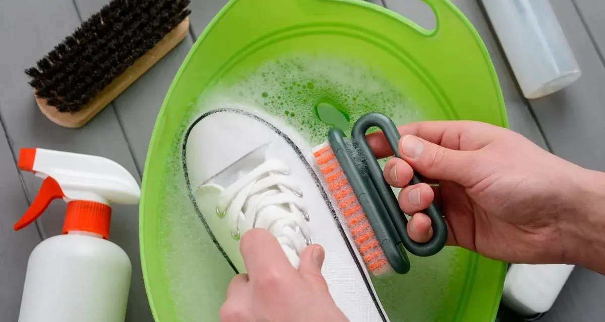 Come lavare le Superga bianche: una scarpa immersa in acqua saponata, mentre una mano la strofina con una spazzola.