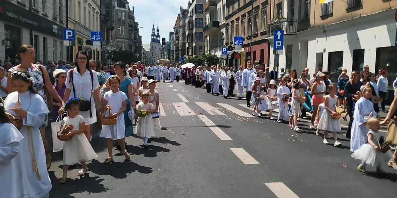 Procesja dzieci w białych strojach, niosących koszyki, idzie ulicą. W tle widać budynki i ludzi, co może symbolizować sekularyzację społeczeństwa.