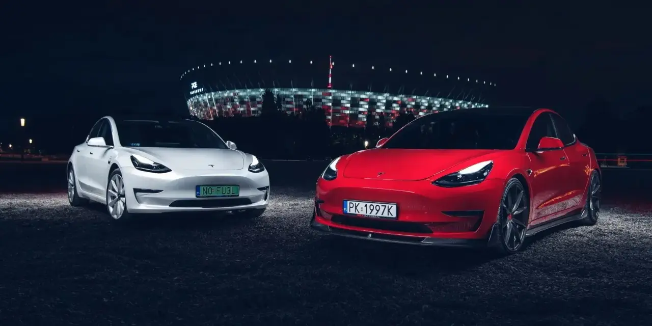 Dwa modele Tesla, biały i czerwony, zaparkowane nocą przed oświetlonym stadionem.