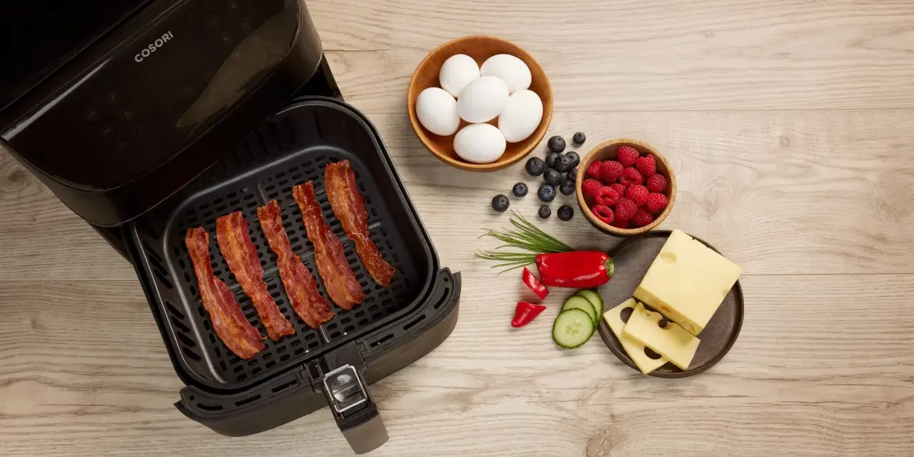 Knuspriger Bacon im Airfryer, daneben Eier, Beeren, Käse und Gemüse für ein perfektes Frühstück.